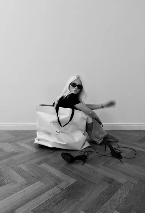 Black-and-white photo of Louisa Reiter sitting on a herringbone floor beside a large paper shopping bag and a pair of heels